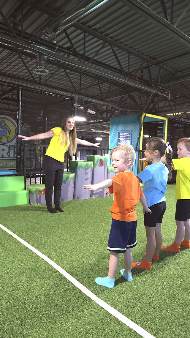 Preschoolers standing in a line on the indoor turf, arms outstretched, following their teacher who leads the exercise